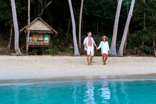 diverse couple of Asian woman and European man walking on the beach at sunset, on the background a wooden bamboo hut bungalow. a young couple of men and woman on a tropical Island in Thailand