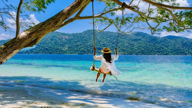 woman swinging around under a tree at a swing on the beach of Koh Lipe Island Southern Thailand, turqouse colored ocean and white sandy beach, Asian female with a summer hat on a swing, rear view