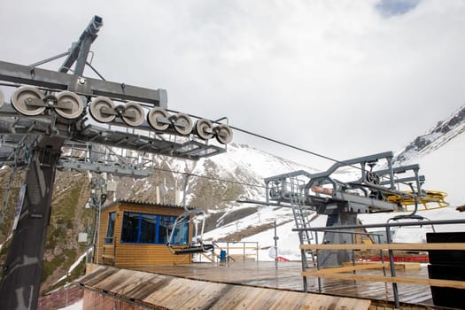 A yellow and gray ski lift wheel and tower with a blue sky and snow-capped mountains in the background. The ski lift is surrounded by red and white striped safety barriers.