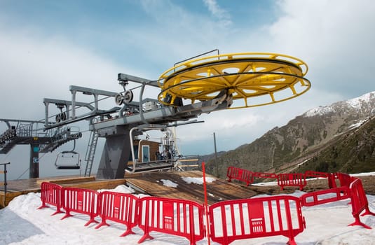 A yellow and gray ski lift wheel and tower with a blue sky and snow-capped mountains in the background. The ski lift is surrounded by red and white striped safety barriers.