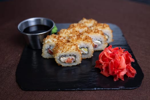 Deep-fried sushi roll on black stone plate with soy sauce, wasabi. Made with rice, seaweed, vegetables, shrimp tempura.