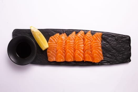 Top view of a delicious and healthy meal of fresh salmon fillet sliced into thin strips and served on a black stone plate with soy sauce and a lemon wedge. The plate is isolated on a white background.