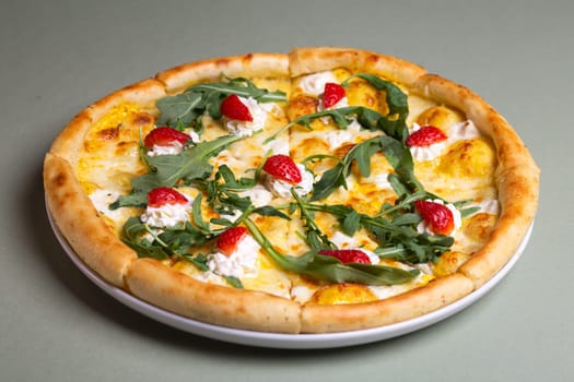 A delicious pizza with strawberries, cream cheese, and arugula on a white plate. The pizza is isolated on a green background. The pizza is perfect for a summer party or a picnic.