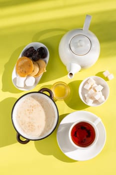 A cozy breakfast setup featuring oatmeal with milk, tea in a white cup on saucer, honey in a glass bowl, and bread slices on a yellow background. Shadows add depth and warmth to the scene.