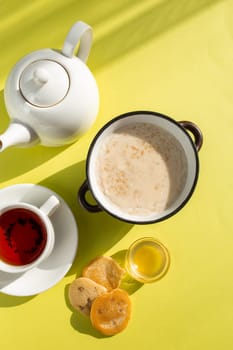 A cozy breakfast setup featuring oatmeal with milk, tea in a white cup on saucer, honey in a glass bowl, and bread slices on a yellow background. Shadows add depth and warmth to the scene.