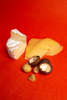 An exquisite assortment of brie, cheddar, and gouda cheeses elegantly presented on a vibrant red background, accompanied by a scattering of delicious macadamia nuts.