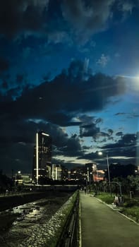 A tall skyscraper stands out among other buildings in the city at night. The sky is dark and cloudy, with a hint of light on the horizon.