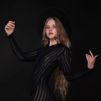 Portrait of blonde young woman looking thoughtfully past camera into distance with her arms elegantly raised to sides. Fashion model wearing black short knitted sheer dress wide-brimmed felt hat