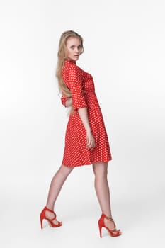 Young blonde female with long hair dressed in summer red polka dot dress, red shoes standing in full length, poses on white background. Caucasian model 21 years old looking over shoulder. Studio shot