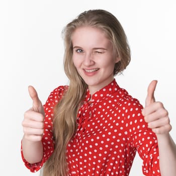 Happy smiling blonde young woman playfully closed one eye and shows thumbs up with both hands. Caucasian female 21 years old dressed in summer red polka dot dress on white background. Studio headshot