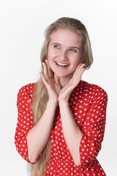 Portrait of blonde young adult woman with positive emotions on her face folded palm under chin and looking away. Playful 21 years old model in red polka dot dress. Studio shot on white background