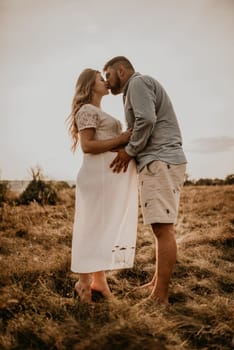 husband hugs pregnant wife keep his hands on big round tummy. Happy family resting in nature hugs kisses in summer at sunset. future mother Caucasian woman in white cotton dress father in shorts