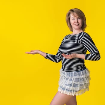 Portrait of curvy blonde woman in mini skirt and striped long sleeve shirt, pointing with open palms to space for future advertising. Woman smiling, looking at camera on yellow background. Copy space