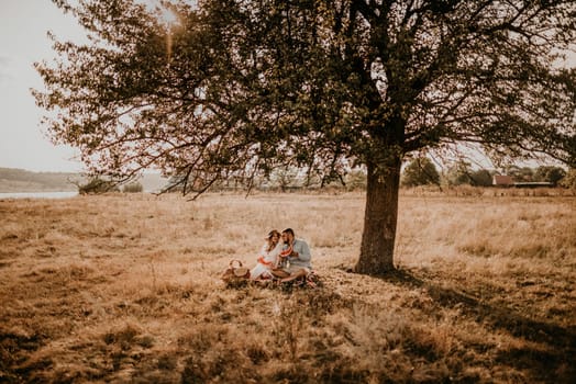 happy European caucasian family with pregnant woman relaxing in nature picnic eating fruit juicy watermelon laugh having fun under tree. expectant mother in hat and dress eating watermelon in summer