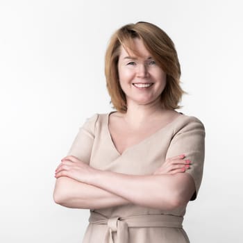 Mature Caucasian woman dressed in casual beige dress looking at camera, standing on white background. Happiness female crossed arms over chest and smiled widely. Part of series