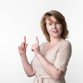 Blonde female 49 year old in beige dress looking at camera with smile, standing on white background. Woman raised index fingers, offering to look at image advertising. Part of series. Copy space