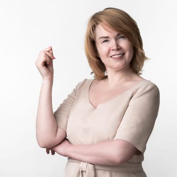 Portrait of smiling happy blonde woman in beige dress looking at camera, standing on white background. Happiness female raised hand and snaps fingers in admiration. Part of series. Copy space