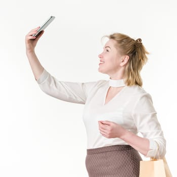Caucasian ethnicity woman taking selfie holding phone up high, holding shopping bags with purchases. 49 years old smiling woman shopper in white blouse and brown skirt. Studio shot on white background