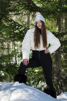 Teenager girl standing in winter forest in snowdrift on background of pine trees. Teen female looking at camera. Full length brunette in knitted hat, white padded bomber jacket, black pants and boots