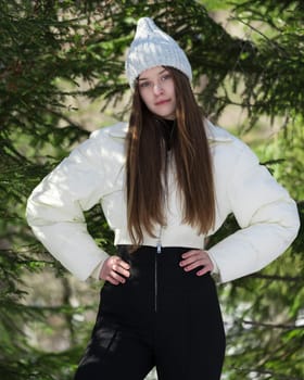 Portrait of beautiful brunette teenager girl arms akimbo, looking at camera. Fashion model standing in winter pine forest. Waist up teen female in hat, white puffer bomber jacket, black pants