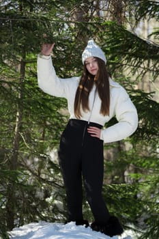 Full length beautiful teenager girl standing in winter pine forest. Brunette teen female looking at camera. Fashion model dressed in knitted hat, white puffer bomber jacket, black pants, winter boots
