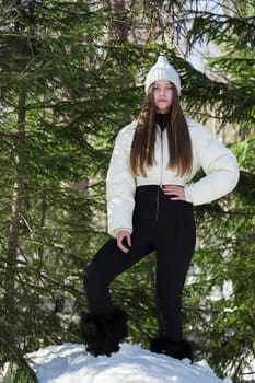 Serene teenager girl standing in winter pine woodland, looking at camera. Full length female fashion model dressed in outdoor clothes - knitted hat, white puffer down jacket, black pants, winter boots