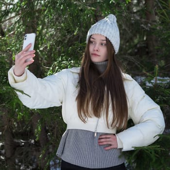 Portrait of brunette teen girl using smartphone making selfie shoots video portrait against backdrop of pine forest. Teen female with long hair, no make-up in knitted hat, white padded down jacket