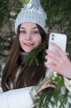 Portrait of cheerful Caucasian teen girl using smartphone taking pictures selfie on pine forest. Happiness and smiling female model generation z dressed in knitted hat, white puffer bomber jacket