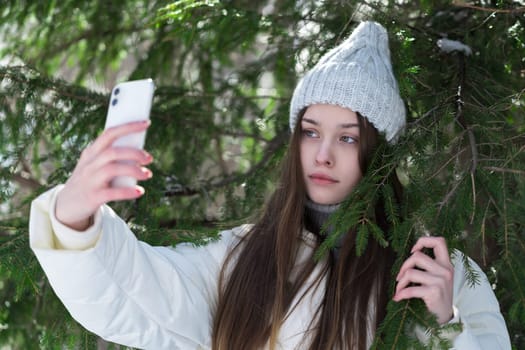 Serious teenager girl using smartphone making video selfie on pine forest. Portrait of 17 year old teen female with long hair, without make-up dressed in knitted hat, white puffer down jacket