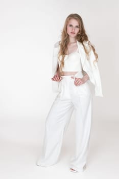 Well dressed Caucasian woman with blonde long wavy hair wearing white suit jacket draped over shoulders, sculpting cupped corset top, pants and sandals. Full length portrait of blondie fashion model