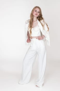 Full length portrait of well dressed woman wearing white suit jacket draped over shoulders, cupped corset top, pants and sandals. Fashion blondie keeps fingers in trouser belt loops, looking at camera
