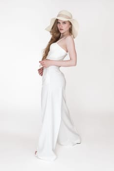 Blonde woman stylishly dressed floppy sun hat, sculpting cupped corset top and trousers stands on white background. Full length side view Caucasian fashion model with wavy long hair looking at camera