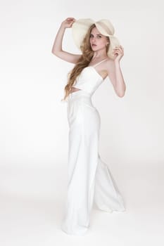 Full length blondie fashion model holding brim of straw hat with both hands and looking at camera. Caucasian woman wearing white sculpting cupped corset top and pants, standing on white background