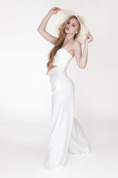 Female fashion model holding brim of floppy sun hat with both hands and looking down. Full length shot fashionable blonde woman wearing white cupped corset top and pants, standing on white background