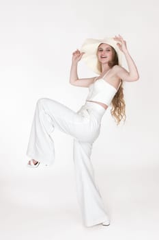 Happiness woman standing on one leg, holding brim of floppy straw hat with both hands, looking away. Full length view of posing woman stylishly dressed sculpting cupped corset top, trousers, sandals