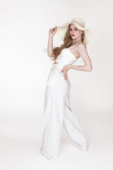 Blondie fashion model holding brim of straw hat with one hand, puts other on waist and looking down. Woman stylishly dressed white sculpting cupped corset top and pants standing on white background