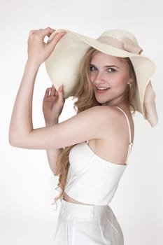 Portrait of fashionable Caucasian woman with raised hands holding brim of straw hat. Headshot smiling young adult female dressed white cupped corset top, pants. Waist up, side view on white background