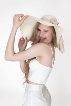 Portrait of beauty woman smiles playfully from ear to ear with one eye closed and winking at camera, looking over shoulder. Model raised hands holding brim of hat, wearing cupped corset top, trousers