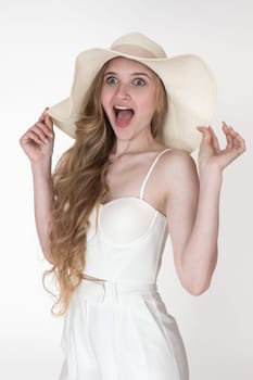 Admiration facial expression of woman stared with mouth open in amazement. Portrait of young adult female model raised arms and holding brim of floppy straw hat with both hands on white background