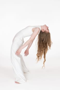 Flexible young blonde woman dancer with eyes closed bending over backwards. Full length, side view of slim barefoot model with long wavy hair wearing white jumpsuit. Studio shot on white background