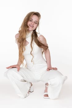 Smiling young woman with eyes closed sitting squatting position put both hands on hips. Playful sensuality blondie model with long wavy hair wearing white long jumpsuit. Studio shot, white background