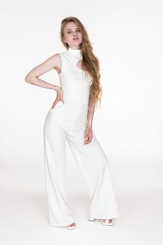 Young adult woman wearing white long jumpsuit office looks looking at camera, standing on white background. Well dressed 21 years old Caucasian fashion model with blonde long hair. Studio shot