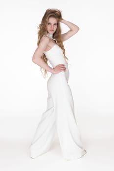 Woman in white long jumpsuit with one hand behind head and other hand on waist standing white background. Young adult fashion model with long hair looking at camera Full length, side view, studio shot