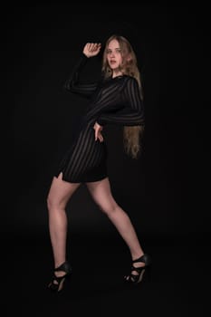 Full length portrait of woman is posing in front of black background. Blonde model with long hair is wearing black short tight knitted transparent dress, wide-brimmed felt hat and high heeled shoes