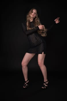 Woman is captured in full length portrait, posing with arms raised to side and gaze fixed downward against black background. She wearing short knitted dress, wide-brimmed felt hat, high-heeled shoes