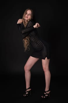 Breathtaking young woman striking confident pose with arms crossed at shoulder height on black background. Blonde with long hair and short knitted black dress, wide-brimmed felt hat, high heeled shoes