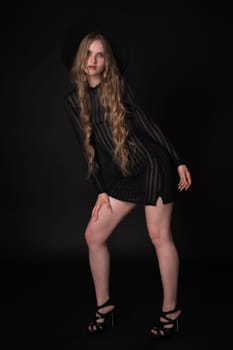 Full length portrait of sexy blonde young woman with long legs on black background. Blonde model with long hair wears black short knitted transparent dress, wide brimmed felt hat, high heels shoes