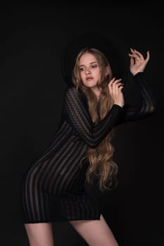 Blonde model posing against black background, with arms raised and wide-brimmed felt hat on head. Beautiful woman in black short knitted translucent tight dress make her look so fashionable and chic