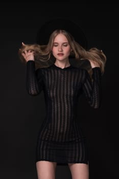 Portrait slim young blonde woman with closed eyes and hands in motion straightening long hair. Model in short tight knitted translucent dress and wide-brimmed felt hat. Black background adds to drama