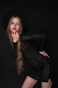 Portrait of young blonde woman striking graceful pose, looking coyly to side with finger to lips, one hand on waist. Model in short black knitted see-through tight dress, felt hat on black background